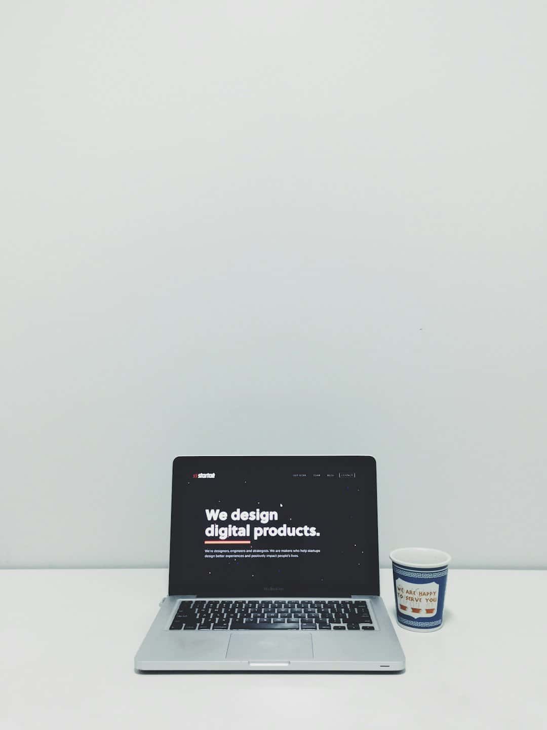 Srebrny laptop wyświetla napis „Projektujemy produkty cyfrowe” na białym biurku.